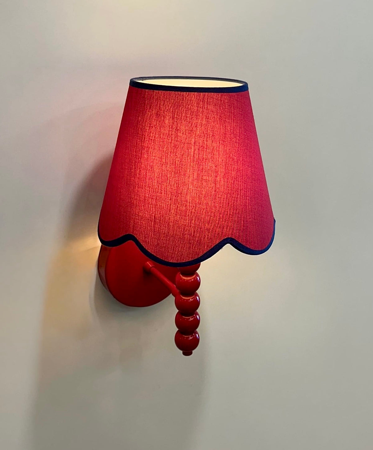PROMIN red wall sconce lit, red fabric shade with scalloped hem casting warm light on the wall